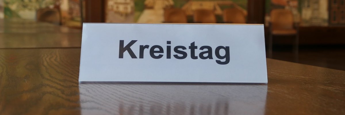 Das Schild, auf das "Kreistag" gedruckt ist, steht auf einem braunen Tisch, von dem nur die Tischplatte zu sehen ist. Im Hintergrund ist verschwommen die Wandmalerei des Großen Sitzungsaales.