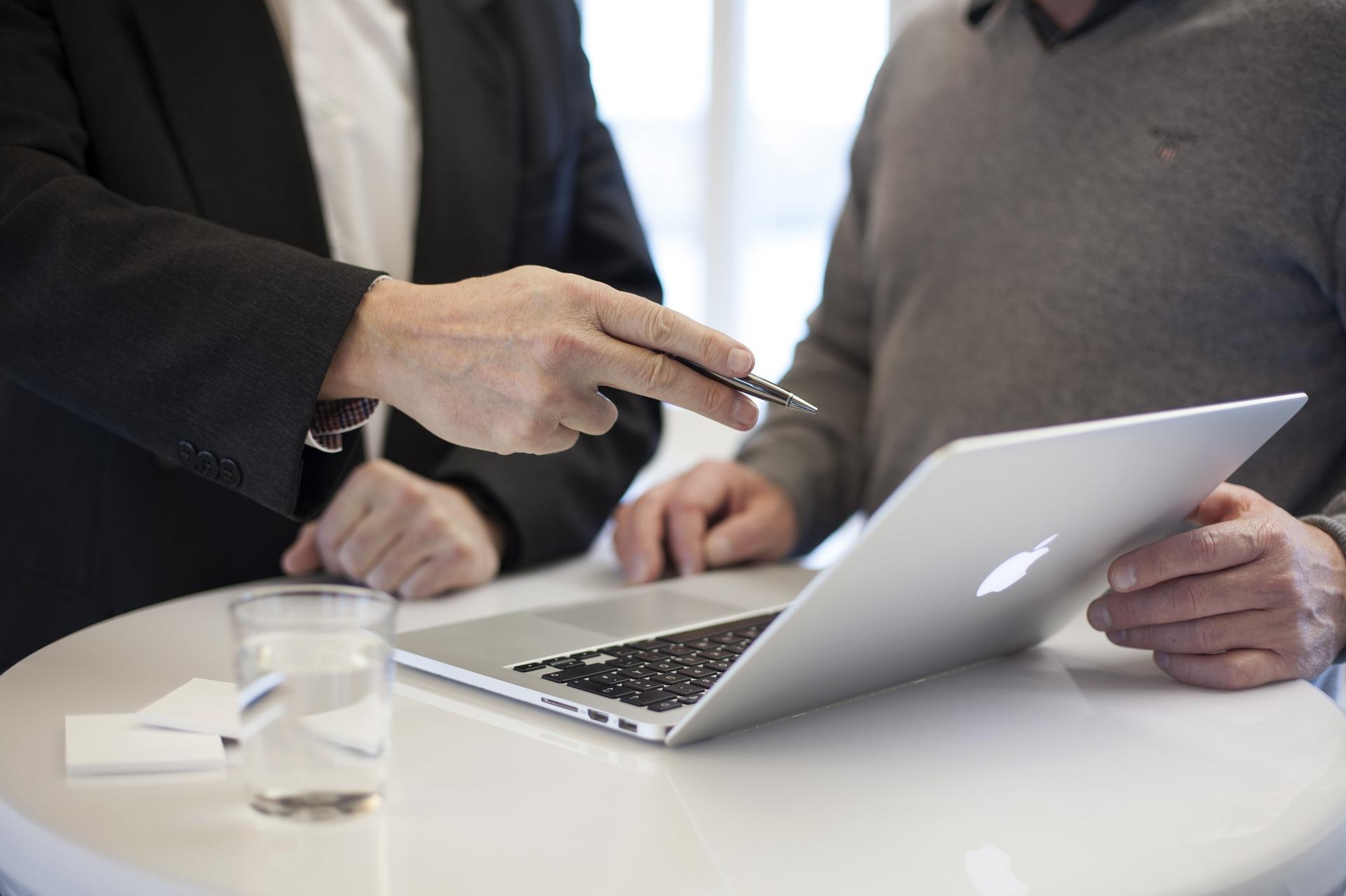 Besprechung und Erklärung am Laptop Auf einem Tisch steht ein Laptop-Computer. Dahinter sind zwei Menschen. Ein Mensch erläutert dem anderen etwas.