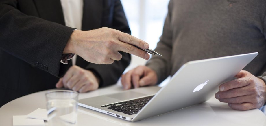 Besprechung und Erklärung am Laptop Auf einem Tisch steht ein Laptop-Computer. Dahinter sind zwei Menschen. Ein Mensch erläutert dem anderen etwas.