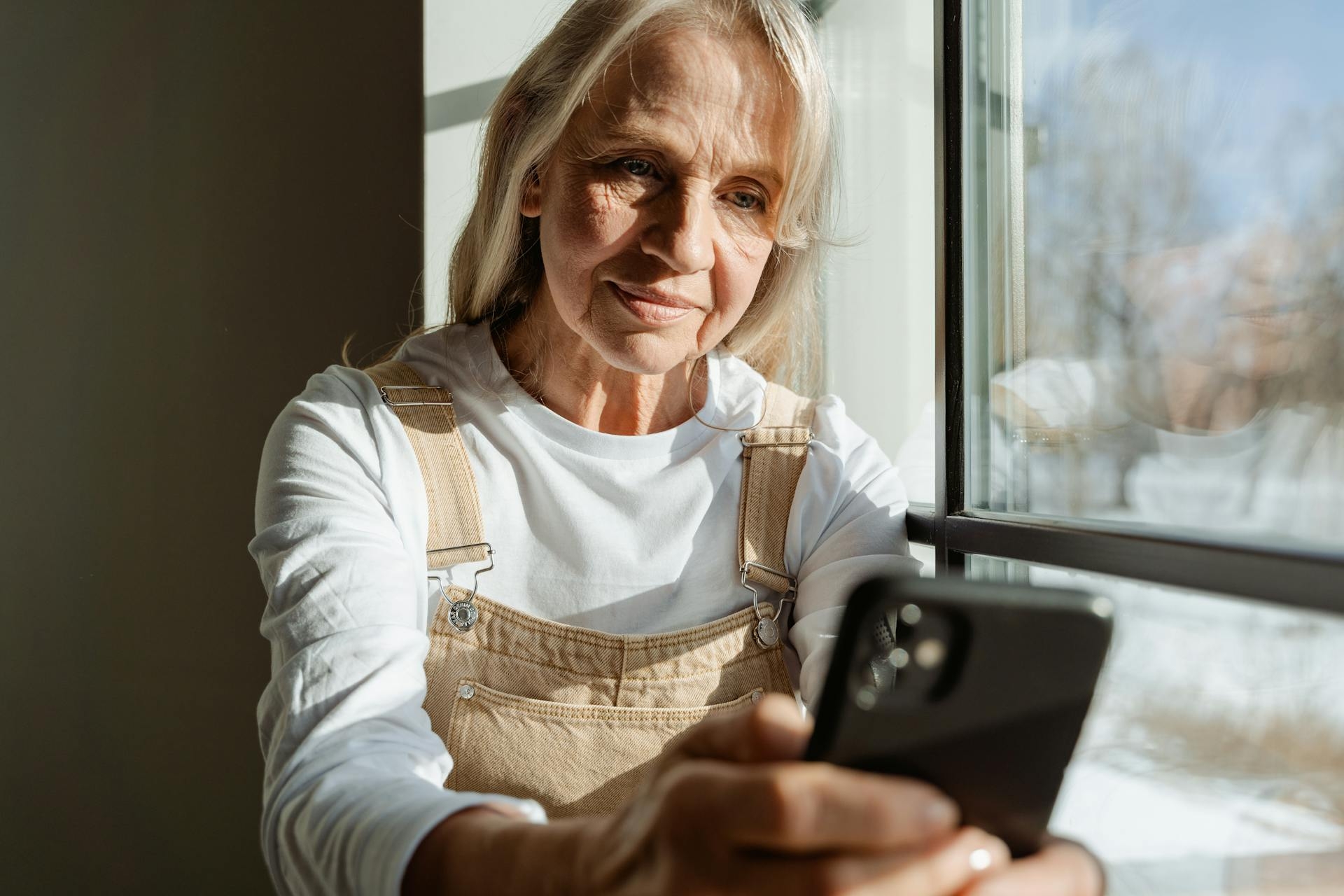 Eine Seniorin sitzt mit Mobiltelefon am Fenster