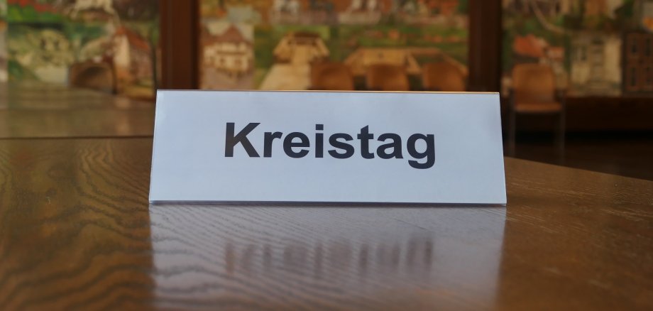 Ein Schild, auf das "Kreistag" gedruckt ist, steht auf einem braunen Tisch, von dem nur die Tischplatte zu sehen ist. Im Hintergrund ist verschwommen die Wandmalerei des Großen Sitzungsaales.