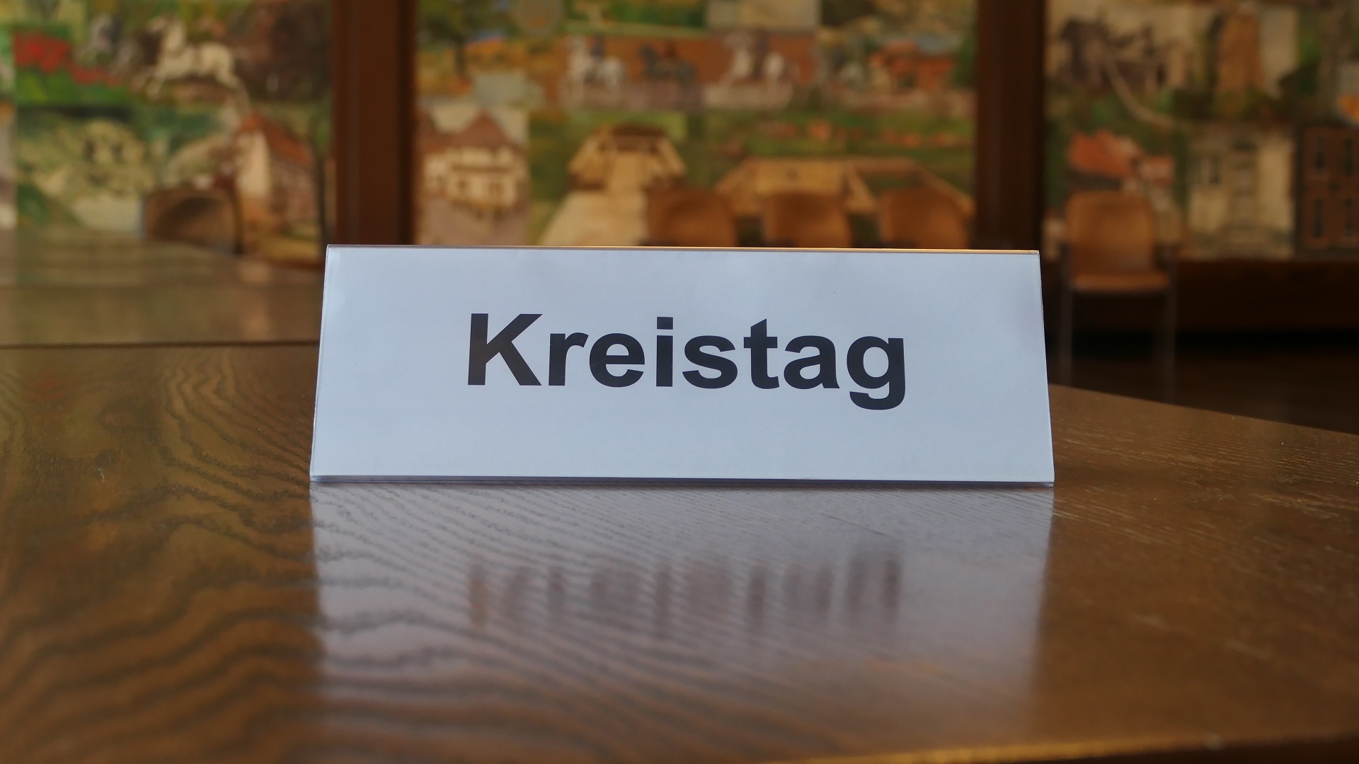 Ein Schild, auf das "Kreistag" gedruckt ist, steht auf einem braunen Tisch, von dem nur die Tischplatte zu sehen ist. Im Hintergrund ist verschwommen die Wandmalerei des Großen Sitzungsaales.