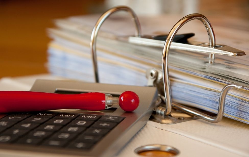 Ein aufgeschlagener Ordner in Nahaufnahme. Auf dem Deckel liegen ein Taschenrechner und ein roter Stift.