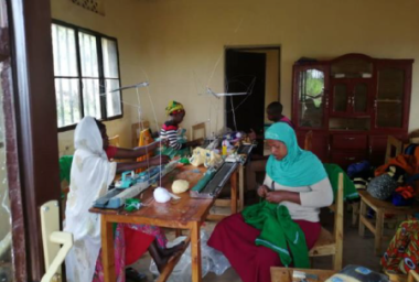 Frauen sitzen an einem langen Tisch, auf dem 4 Strickmaschinen stehen und bedienen diese.