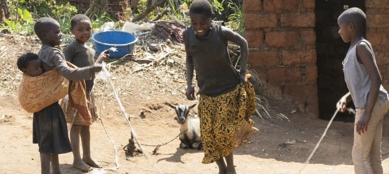 Vier Kinder spielen zusammen. Zwei halten ein Seil, eines springt gerade über das Seil. im Hintergrund liegt eine angebundene Ziege auf dem Boden.