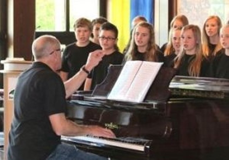 Im Vordergrund spielt ein Mann am Klavier und im Hintergrund stehen Kinder eines Chors mit Mappen und singen.