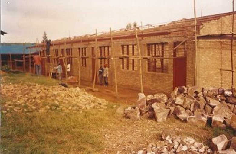 Im Vordergrund liegen große Steine und Schutt. Im Hintergrund steht ein Gebäude aus St4ein ohne Dach mit einem Gerüst aus Holz. Unter dem Gerüst stehen Personen; eine steht auf dem GHerüst.